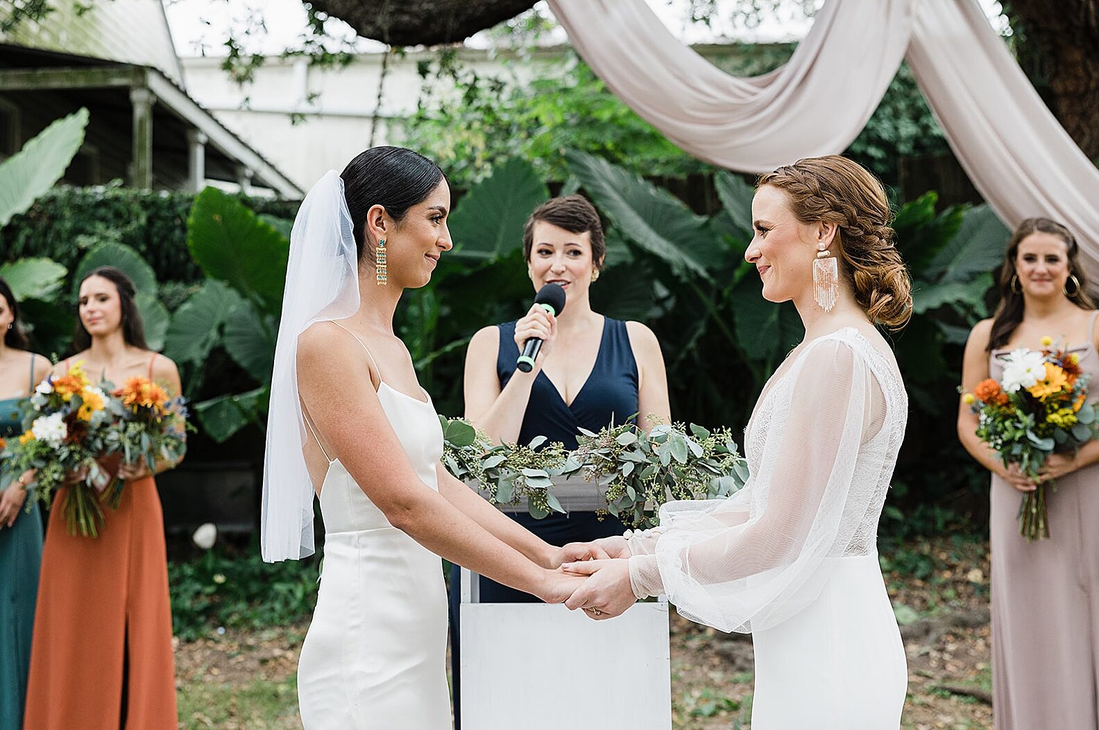 Two brides hold hands during a garden wedding ceremony.