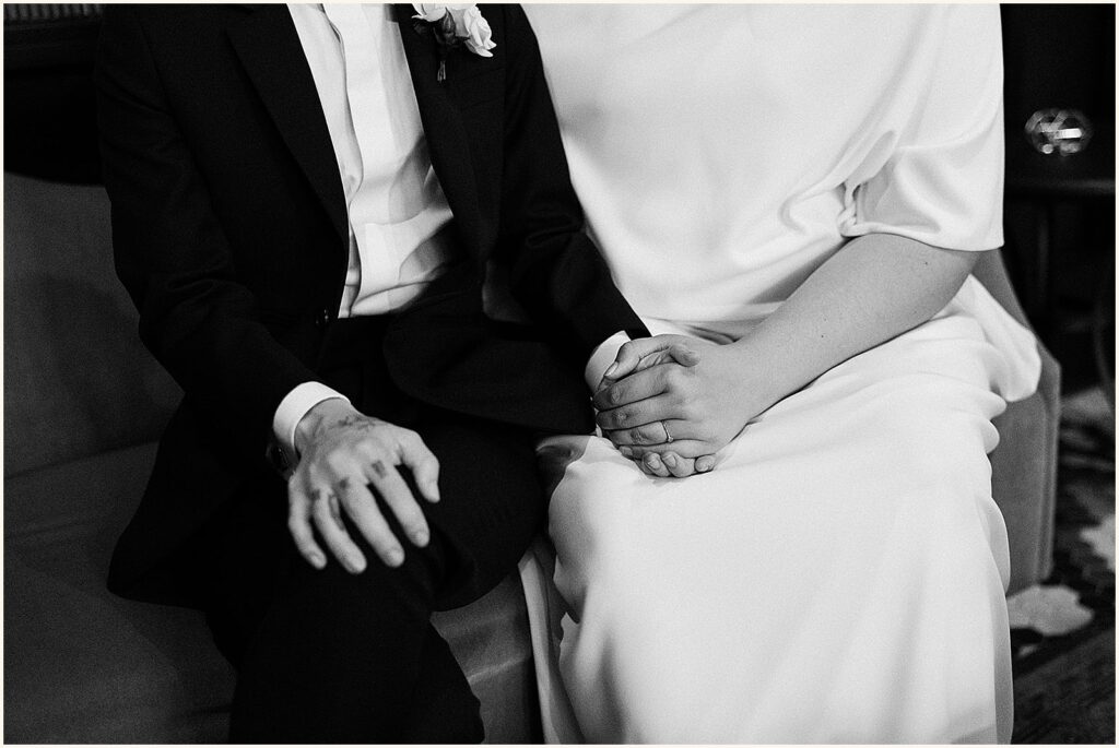 Two marriers in wedding attire hold hands on a sofa.
