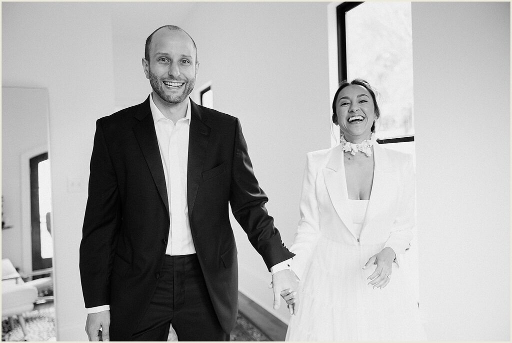 A bride and groom hold hands and laugh in a hallway.