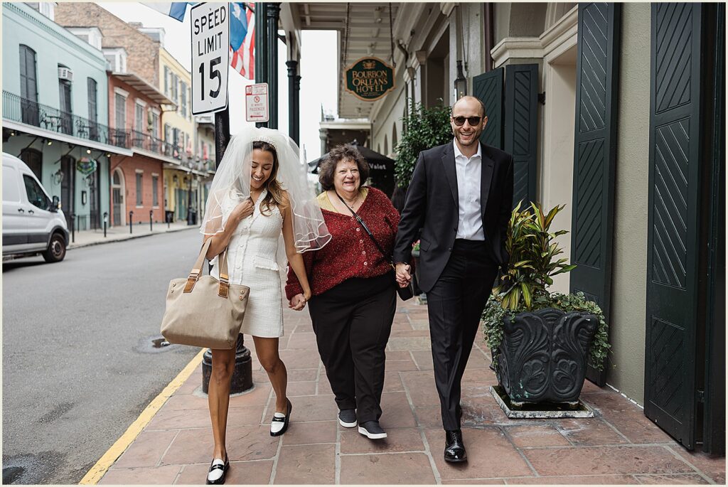 A bride, her mother in law, and a groom walk down a French Quarter sidewalk holding hands.