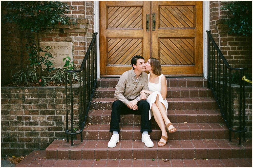 A woman kisses a man on a cheek while they sit on the steps of a Garden District house.