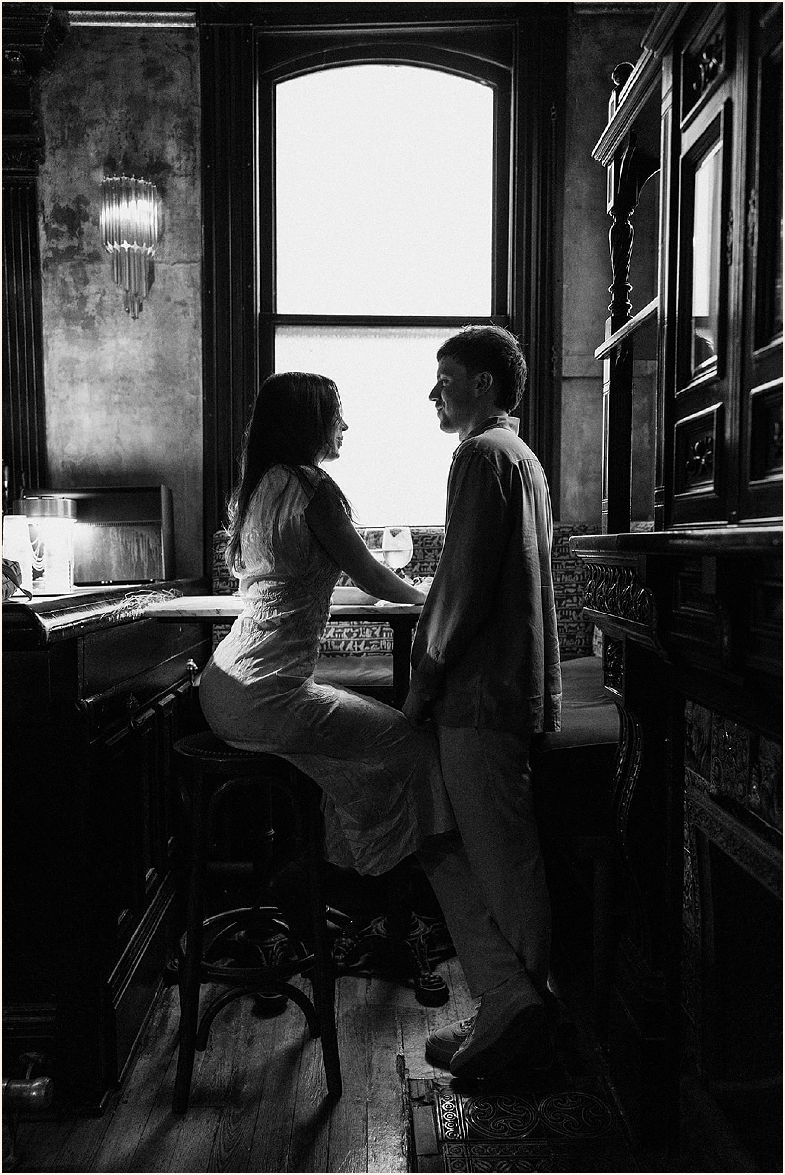 A man and woman stand beside a window in a bar for New Orleans engagement photos.